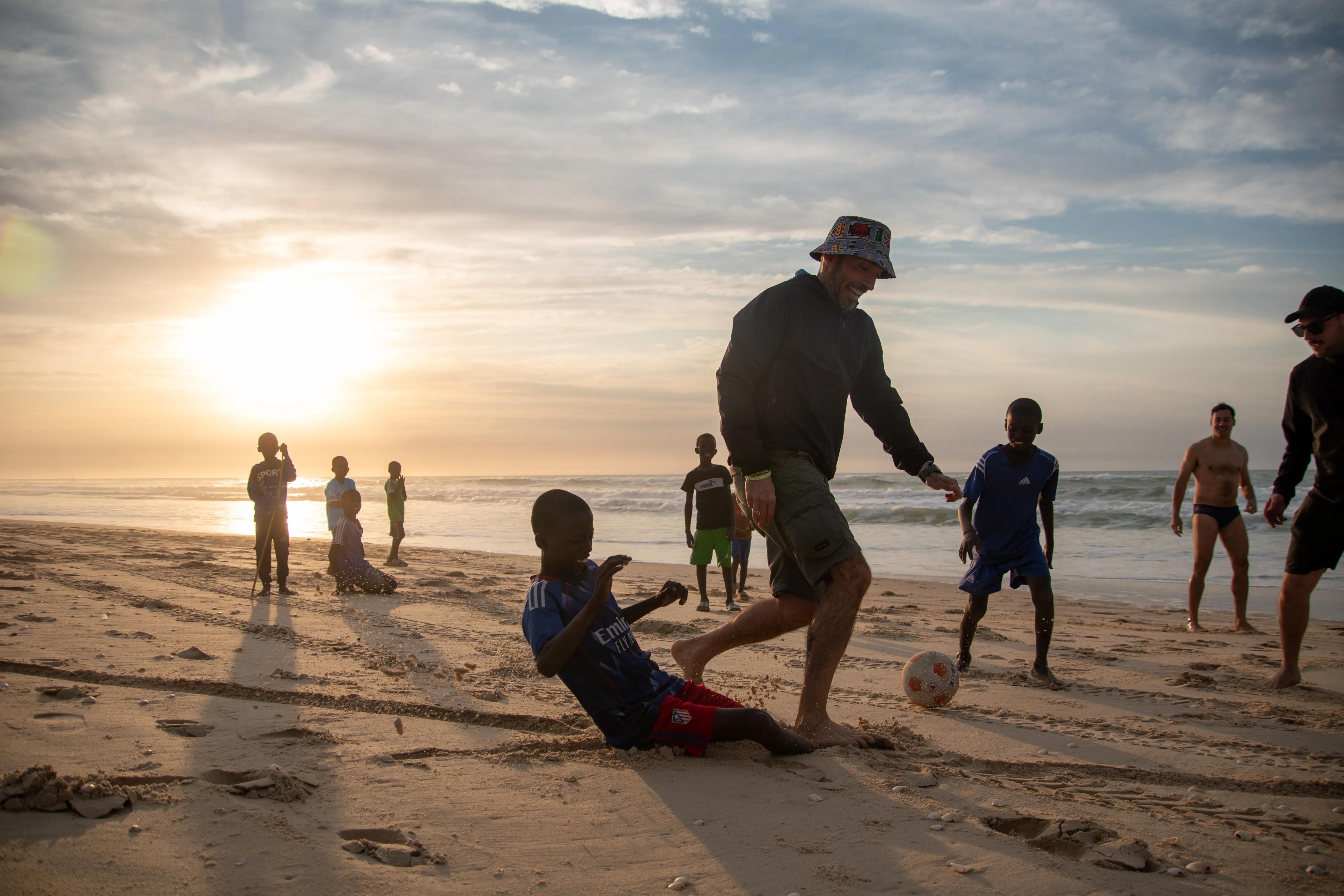 Turisti italiani e bambini senegalesi giocano a calcio sulla spiaggia al tramonto